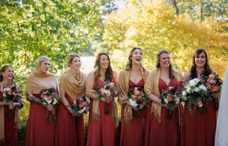 Seven women in matching red dresses and gold shawls holding floral bouquets, standing outdoors with greenery and sunlight in the background.