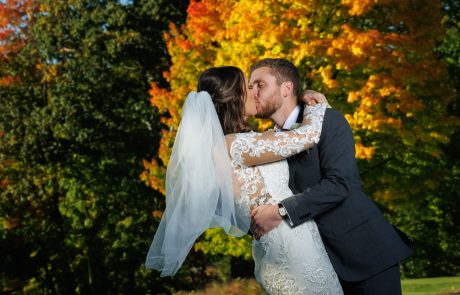 A bride and groom share a kiss in front of vibrant autumn foliage. The bride is wearing a white dress with lace sleeves, and the groom is in a gray suit.