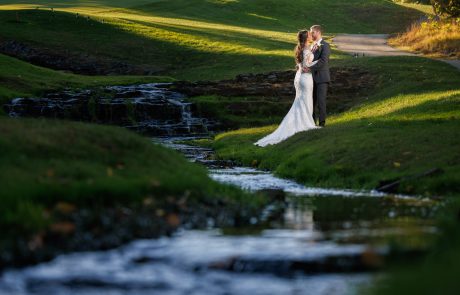 A couple in wedding attire stands on a grassy area near a small stream, embracing each other. The background features a golf course with lush green grass and sunlight filtering through trees.
