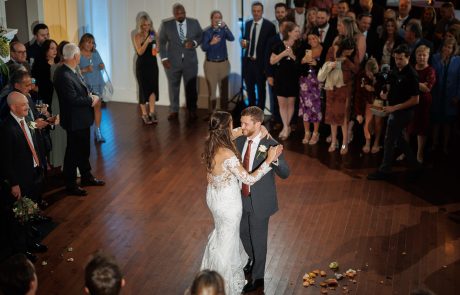 A couple in wedding attire shares their first dance on a wooden floor surrounded by guests in formal attire.