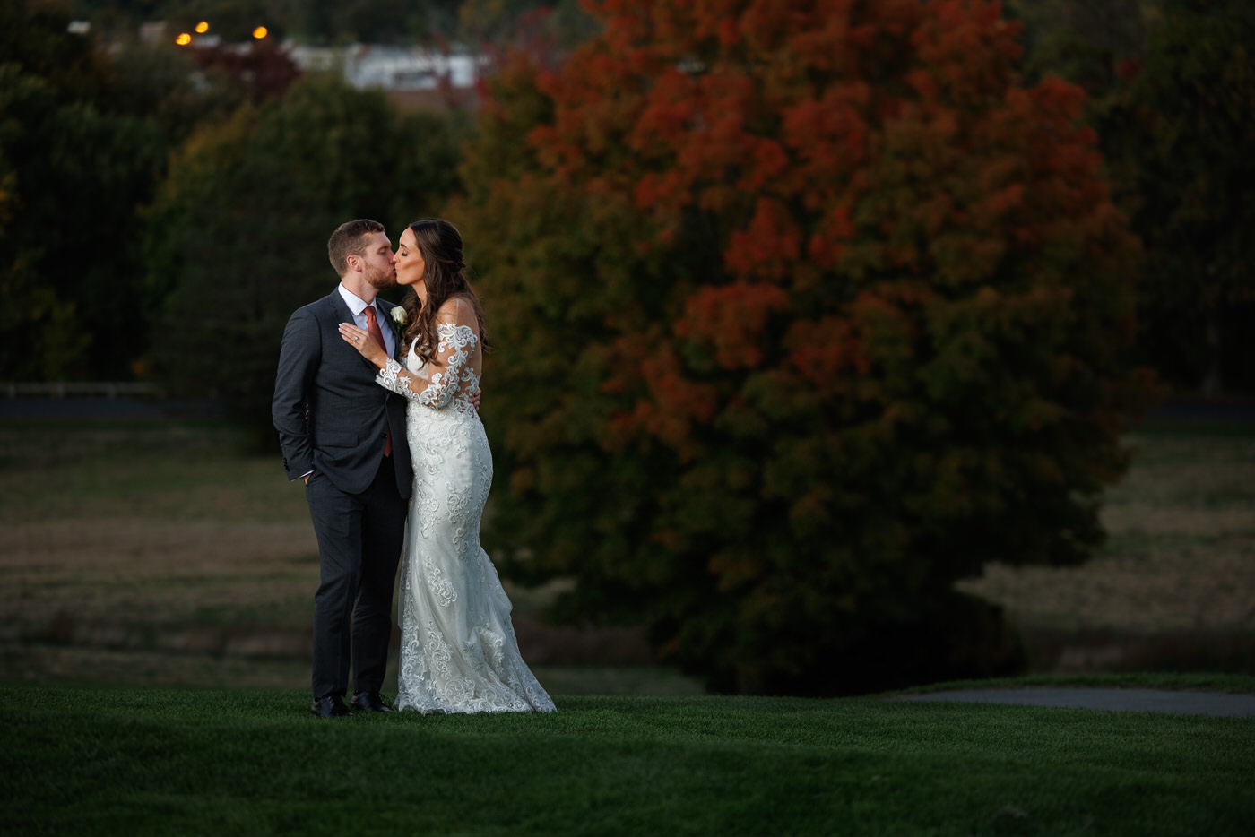 A bride and groom in wedding attire share a kiss outdoors with a large tree showing fall colors in the background.