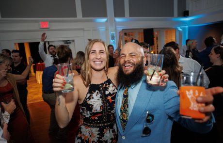 Two people, one in a floral dress and the other in a blue suit, holding drinks and smiling, surrounded by others at a social event indoors.