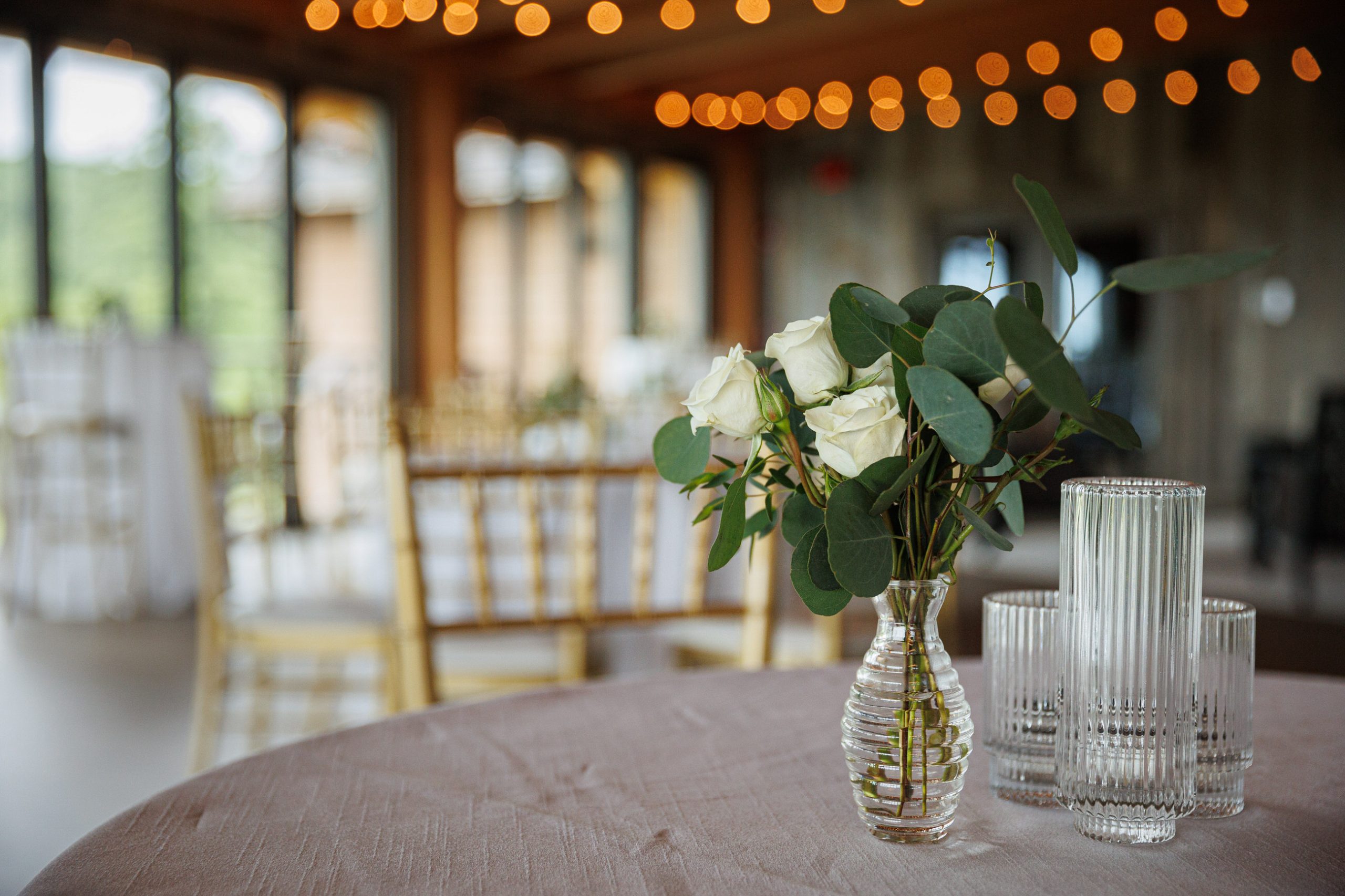 A table with a glass vase containing white roses and greenery, alongside three empty ribbed glass tumblers. The background shows a softly lit room with wooden chairs and strings of lights, setting the perfect ambiance for The Starting Gate Wedding.