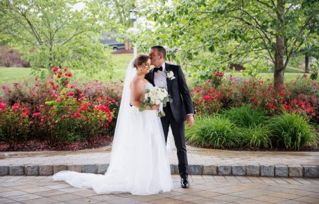 A bride and groom stand on a paved walkway at The Starting Gate Wedding venue, surrounded by blooming flowers and trees. The groom kisses the bride's forehead as she holds a bouquet, both dressed in elegant wedding attire.