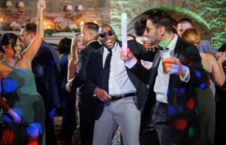 A group of people in formal attire are dancing and laughing at a party, one person holds a glow stick and another holds a drink. Multi-colored lights create a festive atmosphere, just like at The Starting Gate Wedding.