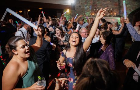 A group of people dance and celebrate at The Starting Gate Wedding, holding drinks and waving light sticks, with expressions of joy and excitement.