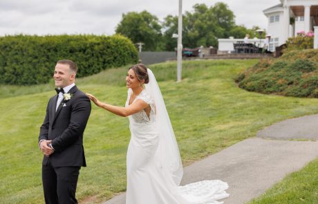 A bride in a white dress and veil touches the shoulder of a groom in a black suit and bow tie, both standing outdoors on a grassy lawn near a white house.