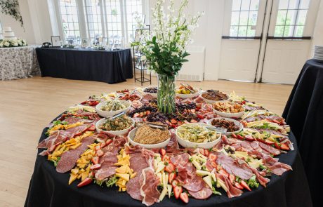 A large round table with various meats, cheeses, and fruits arranged around bowls of salads and dips, adorned with a tall floral centerpiece in the middle.