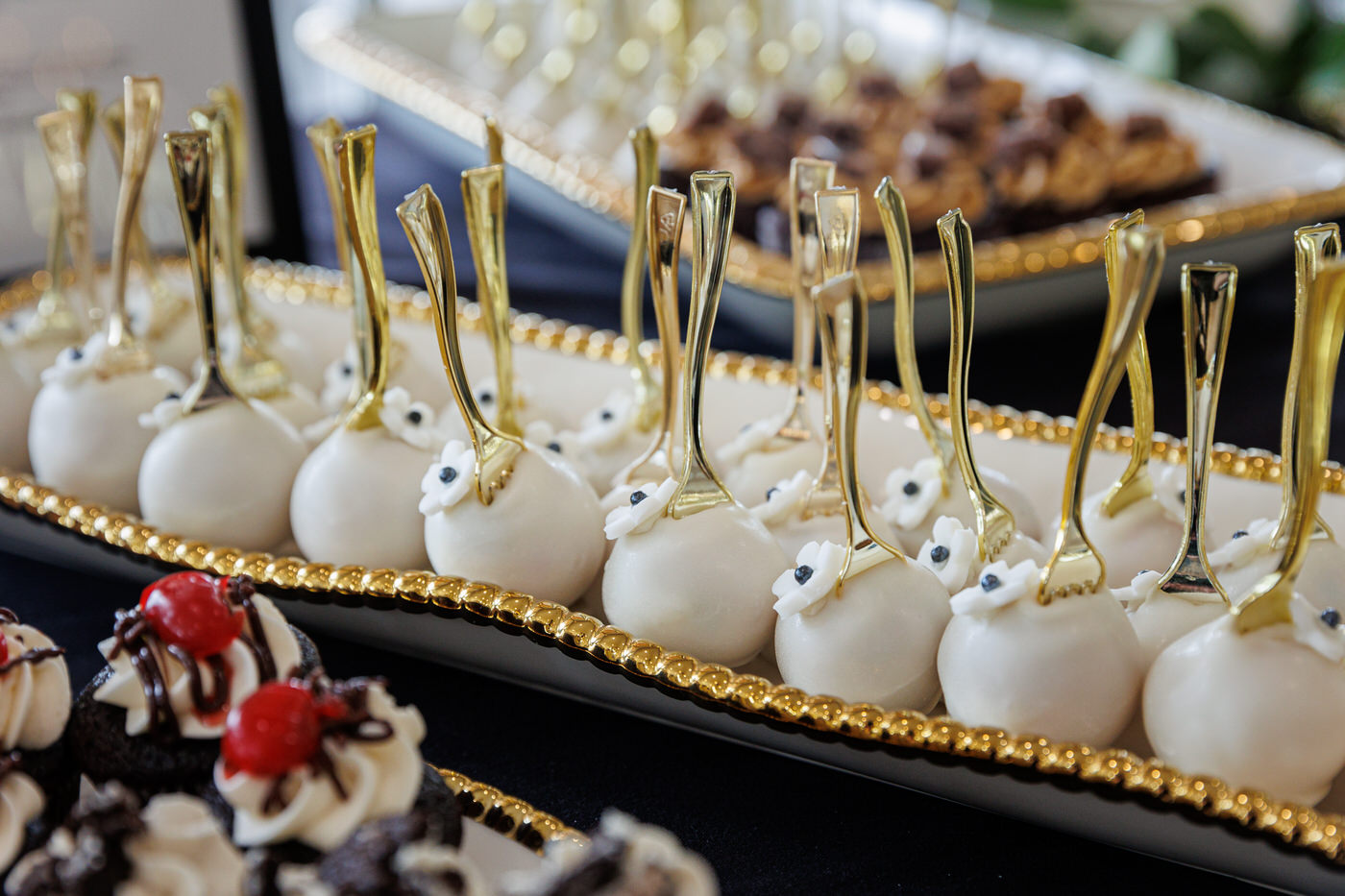 A tray of white chocolate-covered cake pops with golden forks and a cherry-topped chocolate dessert in the foreground.