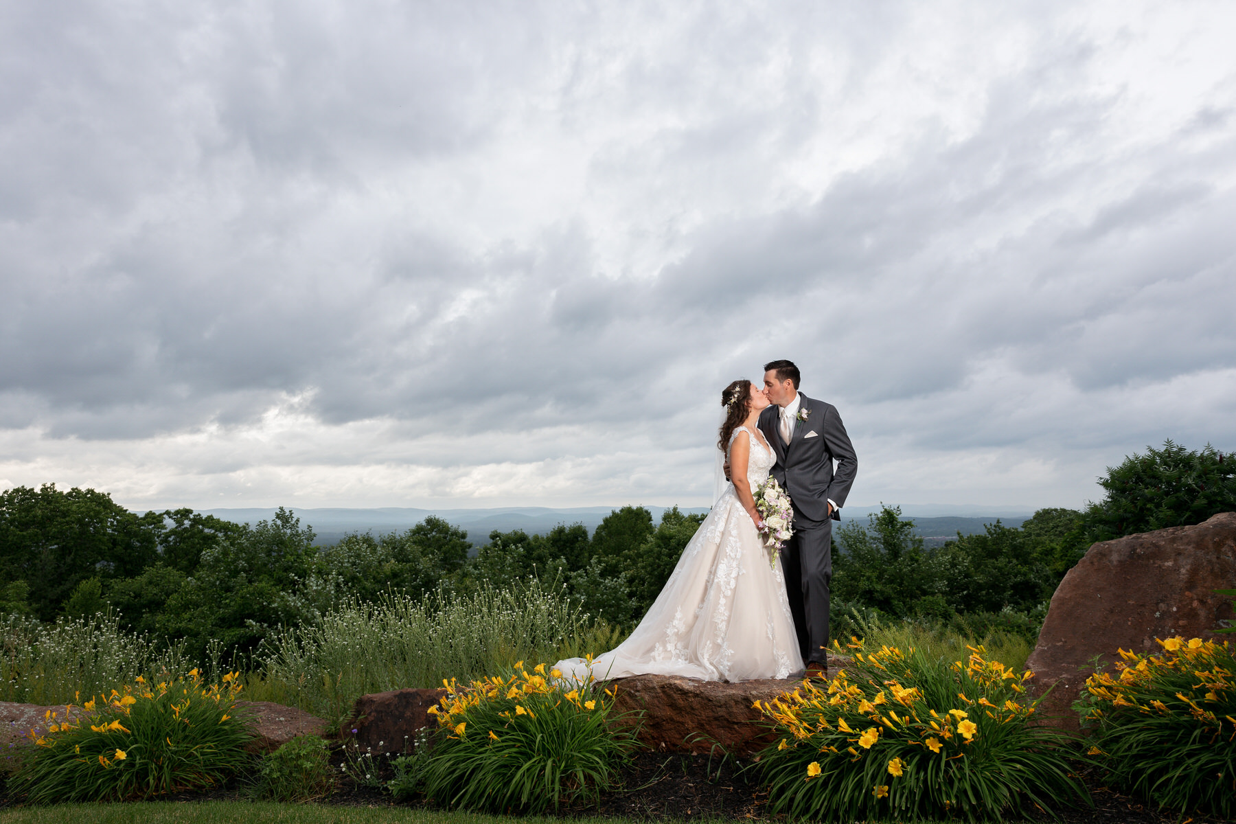 A bride and groom kiss while standing on a rock with a scenic view of the sky and greenery in the background. The bride holds a bouquet, and the groom wears a gray suit. Yellow flowers are in the foreground.