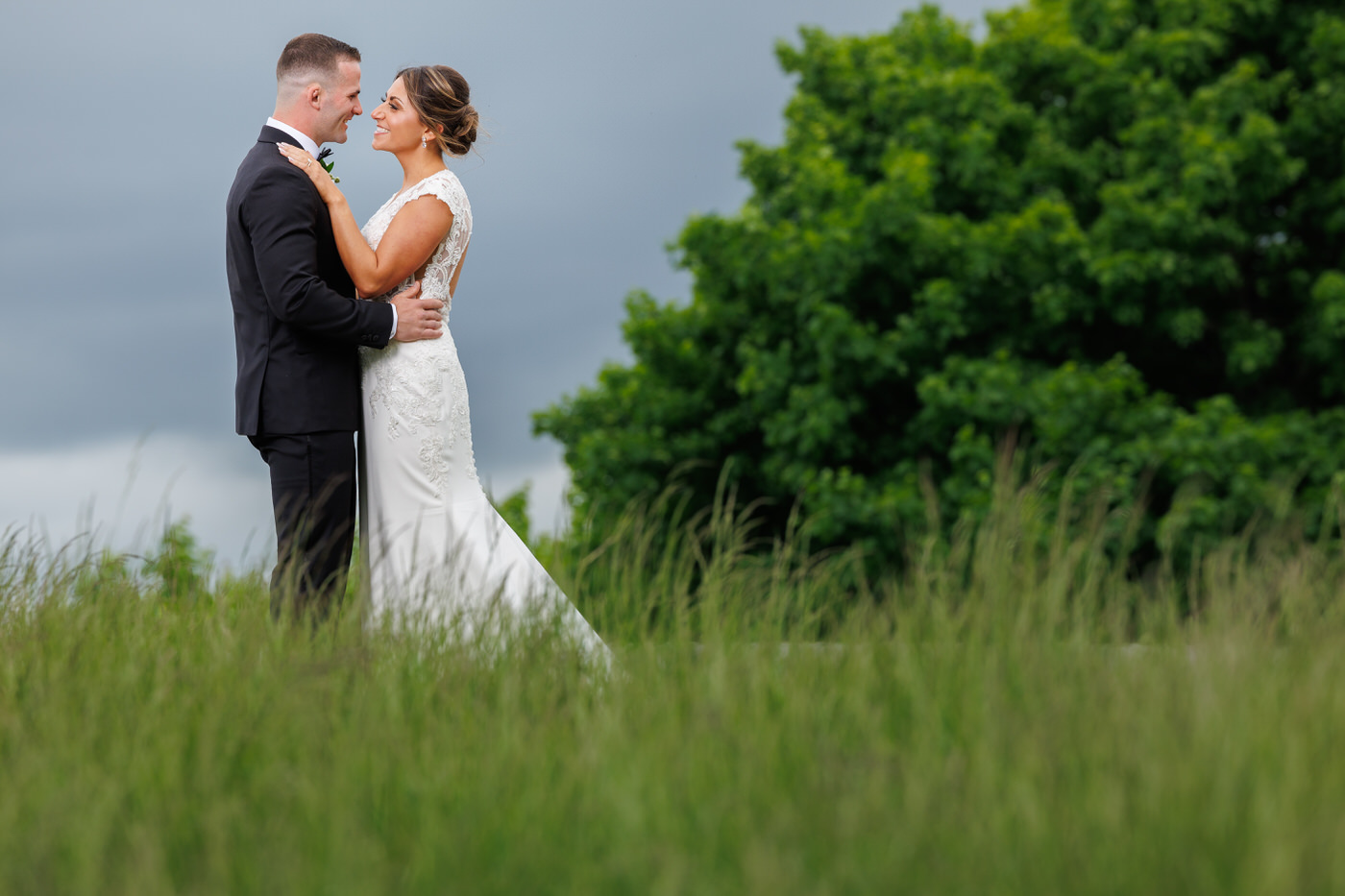 A bride and groom stand facing each other in a grassy field, with the bride wearing a white dress and the groom in a black suit, while trees and a cloudy sky form the background.