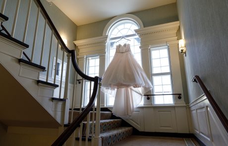 Nicole and Chris-001 A white wedding dress is suspended in front of a large arched window at the landing of a stairway illuminated by wall lights.