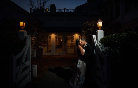 Olivia and Michael-499 A couple in wedding attire shares a kiss at night outside a building with large windows and two lit lanterns.