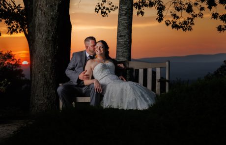 A bride in a white gown and groom in a gray suit sit on a bench under trees at sunset, with the sun setting over distant mountains in the background.