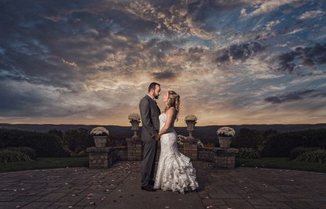 A couple in formal attire stands facing each other, holding hands outdoors, against a backdrop of a dramatic, cloudy sky and distant hills during sunset. Flower petals are scattered on the ground around them.