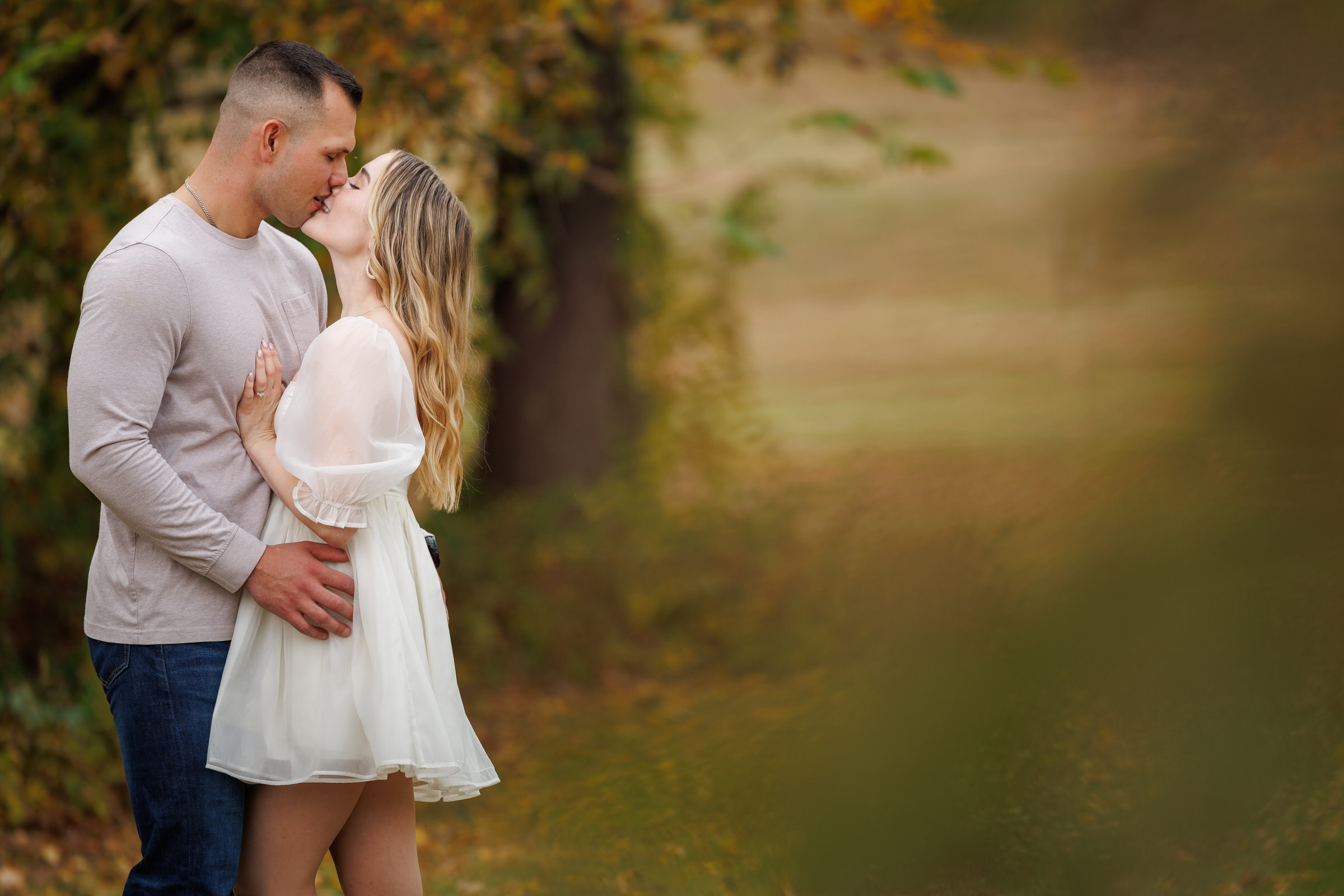 A couple stands outdoors, kissing gently, the woman in a short white dress and the man in a light-colored long-sleeve shirt; perfect engagement photo shoot tips for capturing romance against blurred greenery.