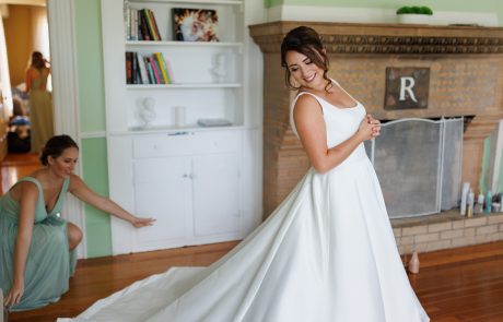 A bride in a white dress stands smiling while an attendant in a green dress adjusts the train of her gown in a room with wooden floors and a fireplace.