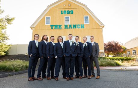 A group of eight men in formal suits stand in front of a yellow building with "1899 THE RANCH" written on it.