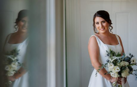 Woman in a white dress holding a bouquet of flowers, standing next to a reflective surface. She is smiling and the background features a light-colored wall.