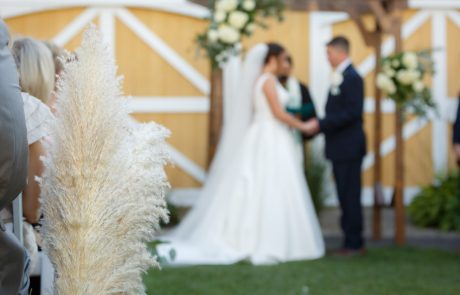 A bride and groom hold hands under a wooden arch decorated with flowers, exchanging vows during an outdoor wedding ceremony. The focus is on a pampas grass arrangement in the foreground.