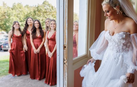 A bride in a white dress and veil stands near a door, smiling at five bridesmaids dressed in matching red gowns, who are standing outside.