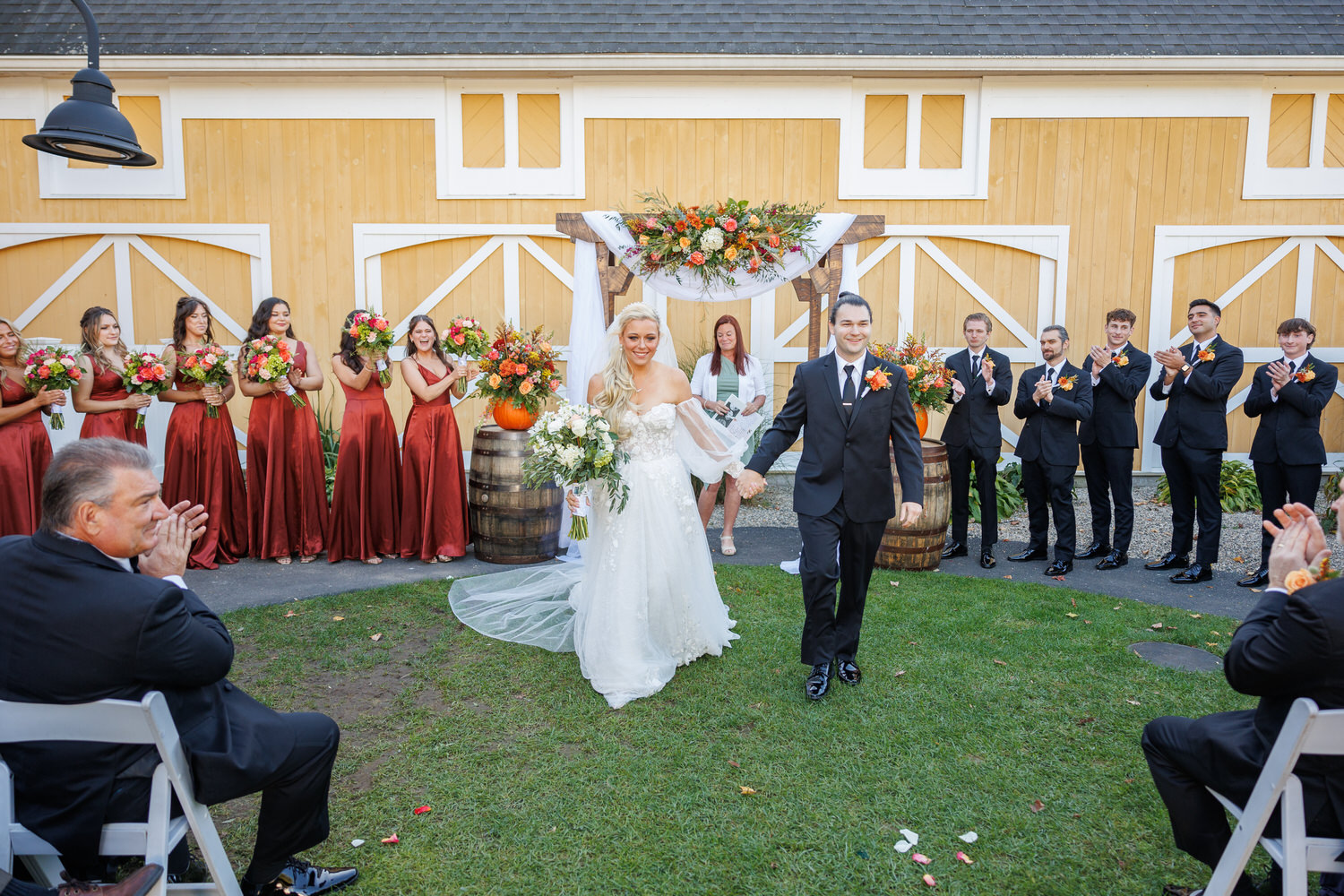 Newlywed couple stands under a floral arch, celebrating with their wedding party and guests in front of a yellow barn.