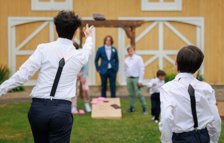 Two people in white shirts and suspenders toss a bean bag towards a cornhole board in a backyard. Other guests in casual attire observe from afar. A barn structure is visible in the background.