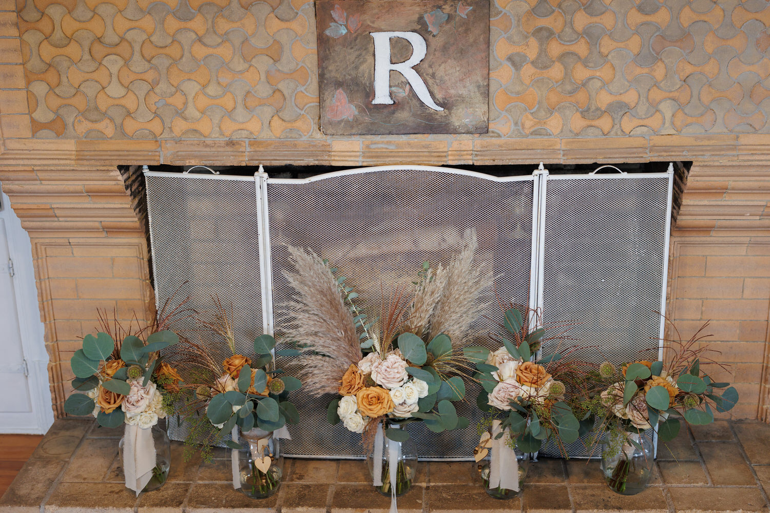 Five floral arrangements with greenery, roses, and pampas grass placed in glass vases in front of a fireplace with a decorative screen and a wall featuring a large letter "R.