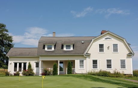 A two-story yellow house with white trim features a front porch, manicured lawn, and flagpole, set against a clear blue sky.