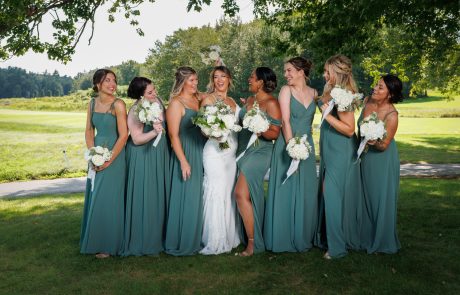 A bride in a white dress stands outside, surrounded by seven bridesmaids in green dresses holding white bouquets. They are smiling and laughing, with greenery and a golf course in the background.