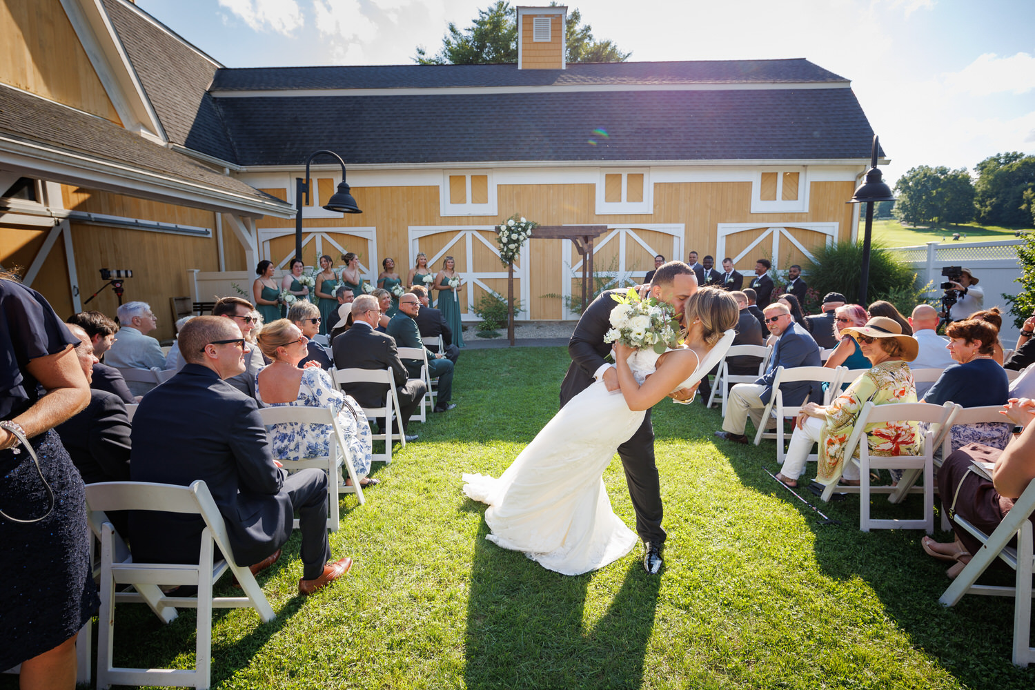 A couple in wedding attire share a kiss at an outdoor ceremony in front of a barn, surrounded by seated guests and their wedding party.