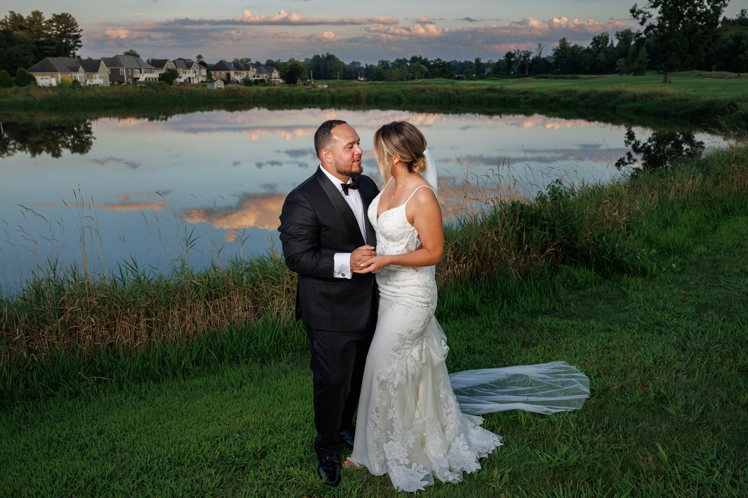 A couple in formal attire stands by a lake, holding hands, with a backdrop of houses, grass, and evening sky reflected in the water.
