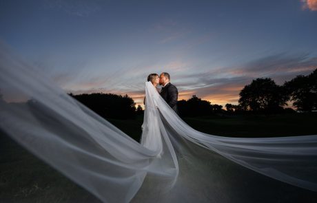 A couple stands in a field at sunset, kissing while the bride's long veil extends dramatically in the foreground.