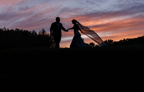 Silhouetted couple holding hands, with the bride's veil flowing in the wind, walking in front of a vibrant sunset sky.
