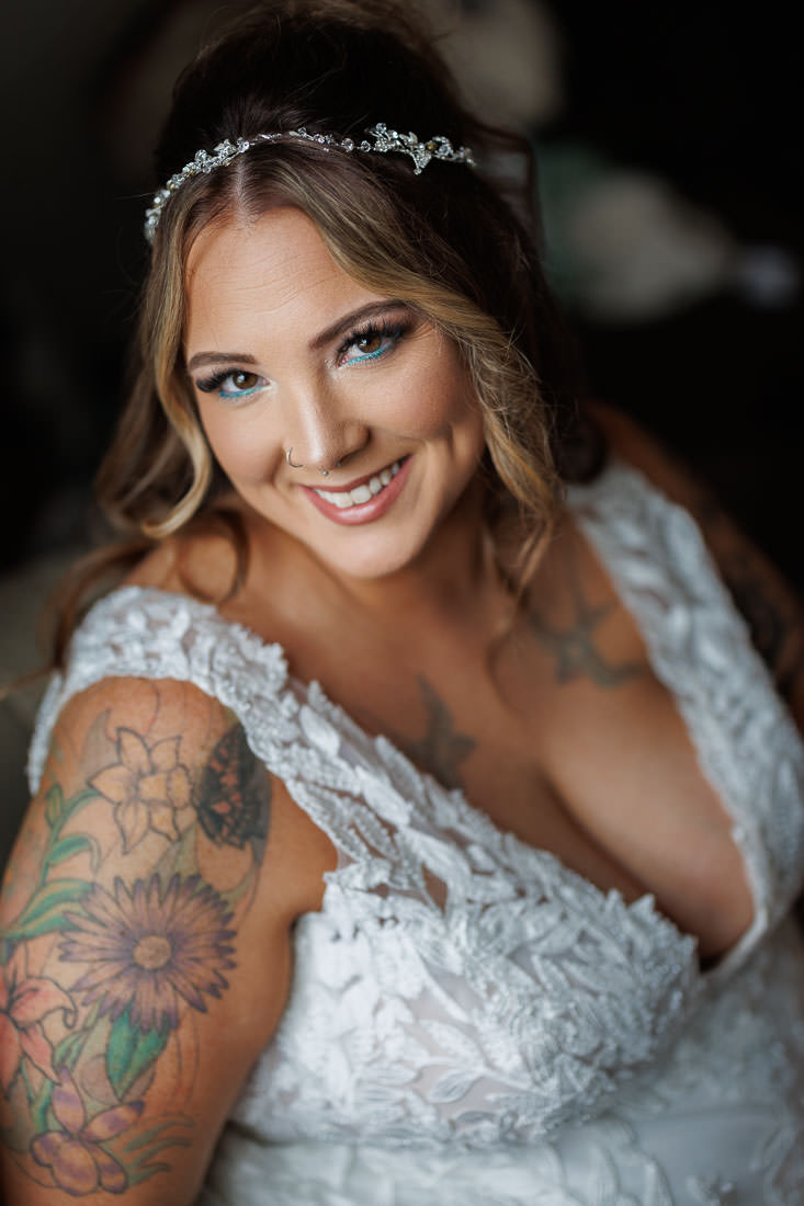 Bliss Farm Wedding-1 A smiling woman in a white dress, adorned with a floral tattoo on her arm and a jeweled headband, capturing the joy of Crestview Country Club wedding photos.
