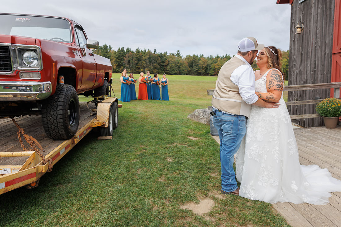 Bliss Farm Wedding-10 A bride and groom share a kiss beside a vintage red truck on a trailer at Crestview Country Club. Bridesmaids in colorful dresses pose nearby, against the backdrop of a wooden building and grassy field, creating picture-perfect wedding photos.