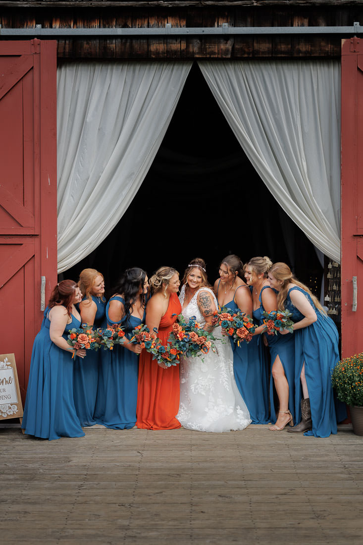 Bliss Farm Wedding-11 A bride in a white dress stands elegantly in the doorway of Crestview Country Club, surrounded by bridesmaids in blue and one in orange, each holding vibrant bouquets.