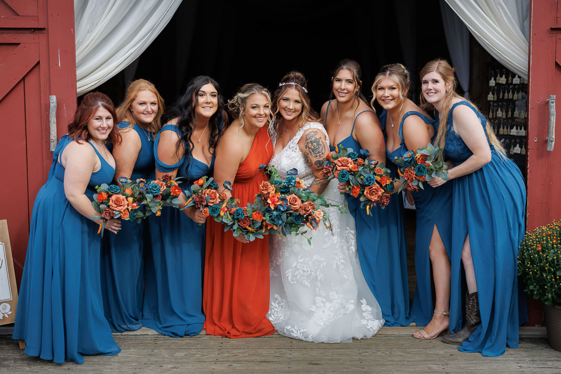 A bride and her seven bridesmaids in gowns pose with bouquets in front of a barn with draped curtains, capturing the essence of a Crestview Country Club wedding.