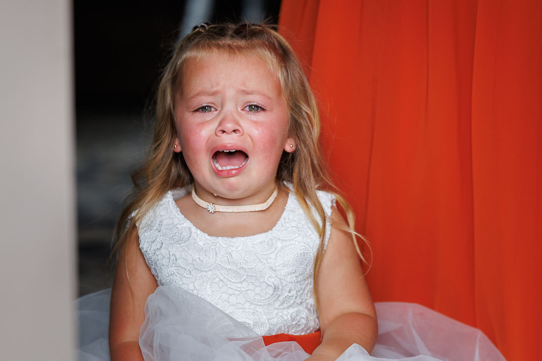 Bliss Farm Wedding-13 A young girl in a white dress is crying, tears streaming down her face, as she stands against the vibrant orange backdrop of a Crestview Country Club wedding.
