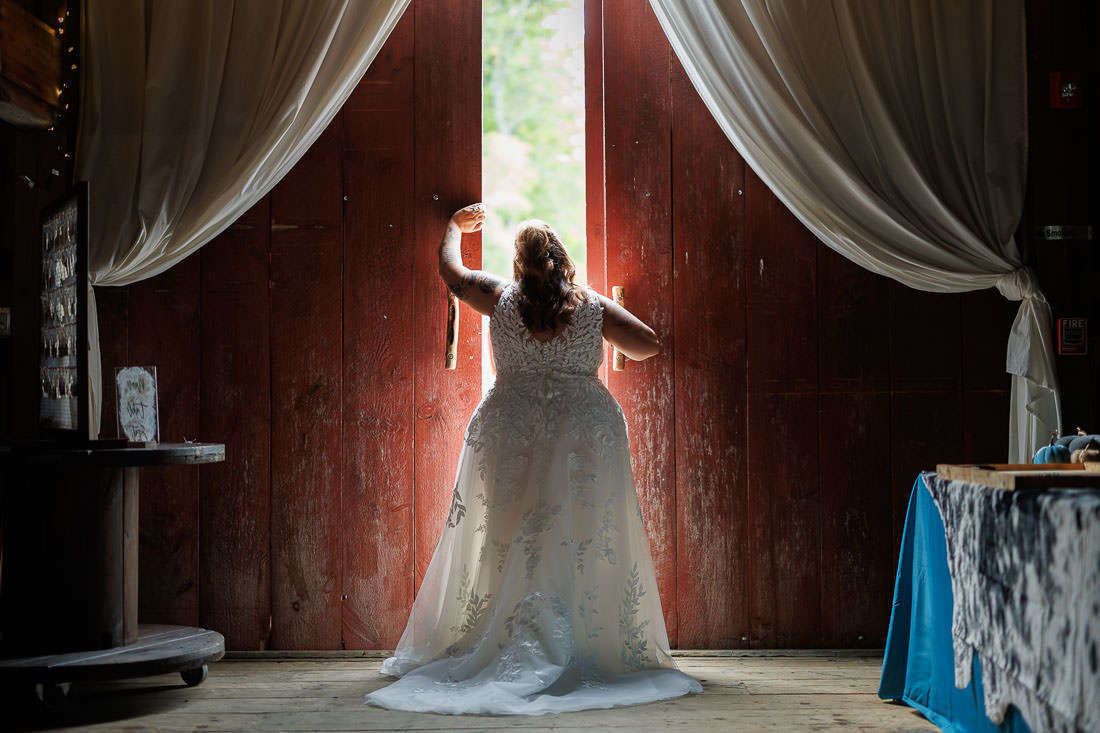 Bliss Farm Wedding-14 A bride stands in a wedding dress at Crestview Country Club, facing large open wooden doors with sheer curtains, gazing outside.