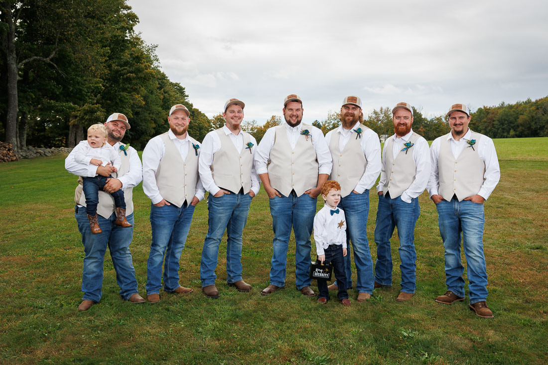 Bliss Farm Wedding-15 A group of eight men in matching attire stand on the lush grass of Crestview Country Club. One holds a child, while another stands beside a child holding a sign. The backdrop of trees and sky captures the perfect setting for wedding photos.