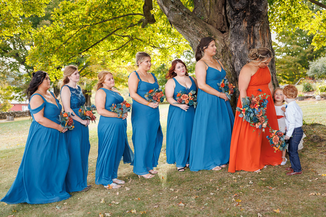 Bliss Farm Wedding-17 A line of bridesmaids in teal dresses stands under a large tree at Crestview Country Club, holding bouquets. They watch a woman in an orange dress chatting with two children, creating picture-perfect wedding photos.