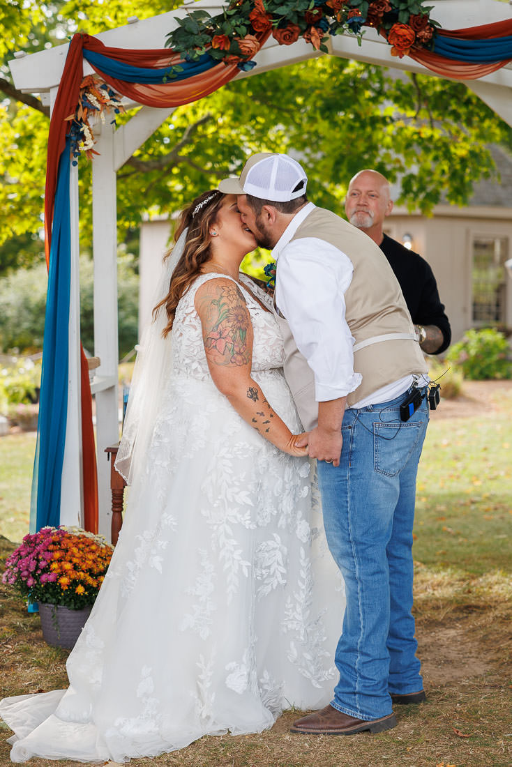Bliss Farm Wedding-18 Bride and groom share a kiss under an outdoor arch adorned with flowers and fabric at Crestview Country Club, holding hands. A man stands behind them, smiling.