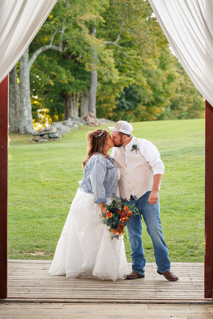 Bliss Farm Wedding-20 A bride in a white dress and denim jacket kisses a groom in a white shirt and jeans under a wooden arch with curtains. They stand before the lush, grassy field at Crestview Country Club, surrounded by trees, capturing a perfect blend of rustic charm in their wedding photos.