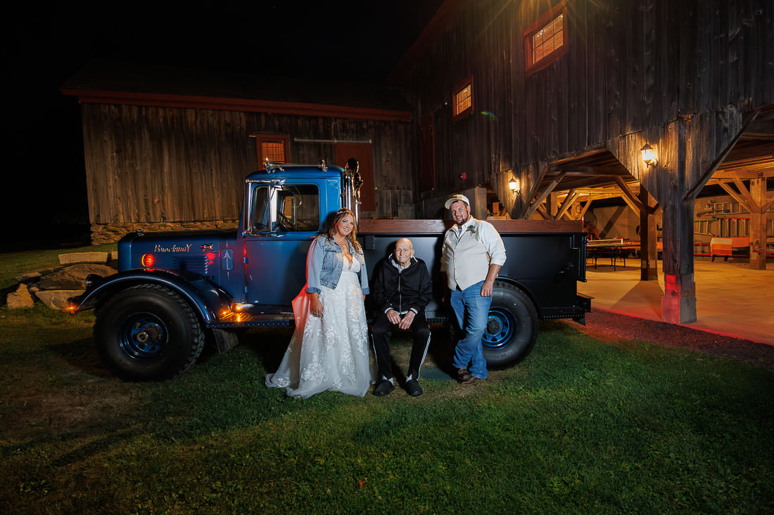 Bliss Farm Wedding-22 Three people pose in front of a blue vintage truck parked by a wooden barn at night, capturing the rustic charm reminiscent of Crestview Country Club wedding photos.