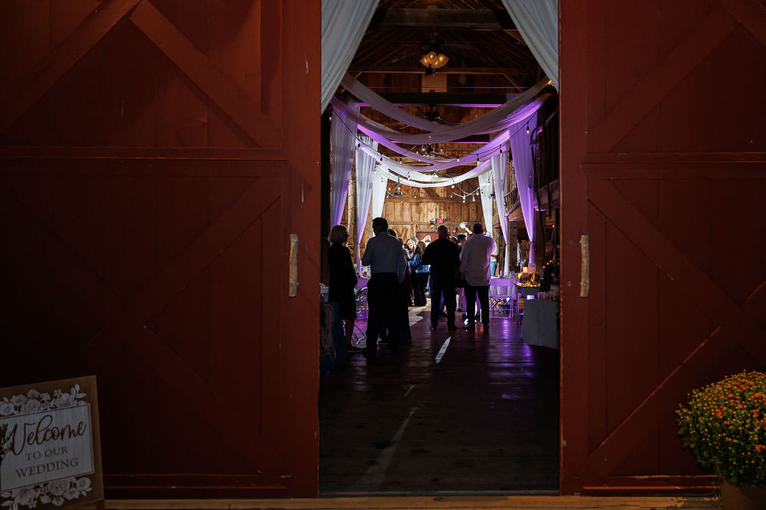 Bliss Farm Wedding-23 Inside the barn at Crestview Country Club, the wedding reception unfolds with draped white fabric and ambient dim lighting. Guests gather around tables, while a "Welcome to Our Wedding" sign warmly greets them at the entrance.