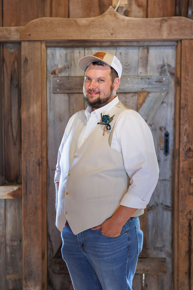 Bliss Farm Wedding-3 Man in a white shirt, vest, and cap stands in front of a wooden door at Crestview Country Club, looking at the camera with one hand in his pocket.