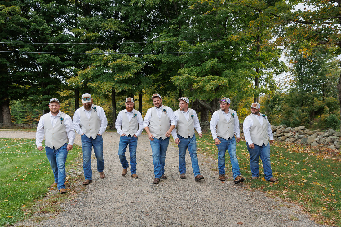 Bliss Farm Wedding-4 Seven men in white vests and jeans walk on a gravel path, surrounded by greenery, capturing the relaxed spirit of Crestview Country Club wedding photos.