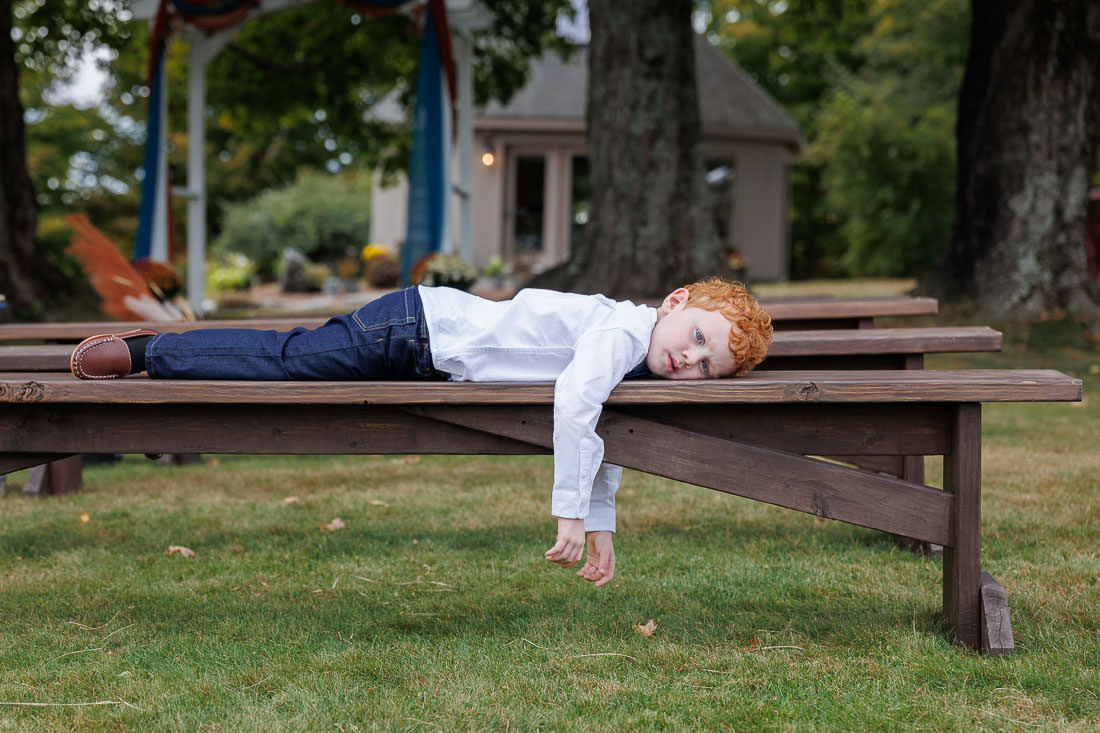 Bliss Farm Wedding-6 A child with red hair is lying on a wooden bench outdoors, appearing sleepy against the backdrop of Crestview Country Club Wedding Photos, with trees and a small building in the background.