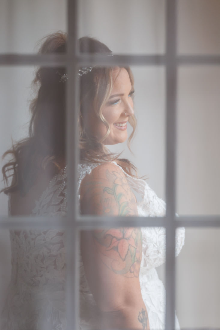 Bliss Farm Wedding-7 Bride in a lace wedding dress with floral tattoos stands behind a frosted window, smiling at her reflection in the Crestview Country Club.